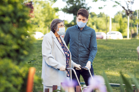 An older woman walking outside using a walking frame. A young man is there to support her. Both are wearing gloves and masks.