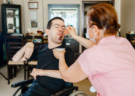 A man in a wheelchair is being assisted to eat a yoghurt by a female PA. They are in his dining room. She is wearing a mask.