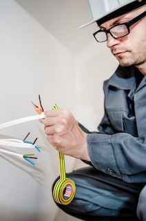 An electrician wearing a white hard hat and blue overalls, is crouching down and putting electrical tape onto the ends of some trunked wiring.
